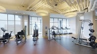 This contemporary fitness center offers a well-appointed workout environment designed to accommodate a variety of exercise needs. The space is defined by its expansive floor-to-ceiling windows, which flood the interior with natural light and frame sweeping views of the surrounding urban skyline, creating an energizing atmosphere for training.

The layout is thoughtfully organized into distinct workout zones. A dedicated strength training area features a multi-function cable machine and additional resistance equipment, while a separate cardio zone lines the windowed perimeter with a row of treadmills, providing users with motivating city views during their workout. A flat exercise bench rounds out the free weights and functional training area.

The industrial-style exposed ceiling, complete with visible ductwork and piping painted in a clean white finish, lends the space a modern, urban aesthetic. Large drum pendant light fixtures provide warm, even illumination throughout. The flooring transitions between rubberized athletic surfaces in a warm terracotta tone and gray, offering both comfort and durability underfoot.

Overall, the fitness center strikes a balance between functionality and design, offering a clean, open, and professionally equipped environment suitable for a wide range of fitness routines.