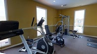 The Derby Estates fitness center is a dedicated, single-floor workout facility designed to support a well-rounded exercise routine. The room features warm, golden-yellow walls that create an energizing atmosphere, complemented by recessed lighting and multiple windows that allow generous amounts of natural light to fill the space. The carpeted flooring provides a comfortable and practical surface throughout the facility. The cardio area is equipped with an elliptical trainer and a stationary recumbent bike, offering low-impact options for cardiovascular exercise. A multi-station cable weight machine anchors the strength training area, providing a versatile range of resistance exercises for various muscle groups. Additional strength equipment is positioned along the perimeter, maximizing the use of the available floor space while keeping the room open and accessible. The windows offer views of the surrounding outdoor environment, adding a pleasant visual connection to the outside while working out. Overall, the Derby Estates fitness center is a practical, well-lit, and thoughtfully arranged space that provides residents with the essential equipment needed for a comprehensive and effective fitness regimen.