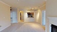 This single-floor space offers an open and functional layout designed to maximize usability. The living area is finished with plush wall-to-wall carpeting in a neutral beige tone, complemented by crisp white baseboards and elegant crown molding that runs along the ceiling perimeter, adding a refined architectural detail to the space.

A striking arched passageway connects the living area to the rest of the unit, adding a classic architectural touch. The kitchen is positioned centrally and features a pass-through counter that opens to the main living space, making it ideal for entertaining. Dark espresso-toned cabinetry provides strong contrast against the light walls, while stainless steel appliances—including a refrigerator, microwave, and range—offer a modern, polished finish.

Track lighting above the kitchen ensures ample task illumination, while recessed lighting throughout maintains a bright and welcoming ambiance. The neutral warm-beige walls create a cohesive palette across the space. Two doorways visible from the main area suggest additional rooms beyond, contributing to a well-organized and practical floor plan overall.