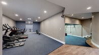 This fitness facility is thoughtfully designed to accommodate a wide range of workout styles within a single, well-organized floor plan. The main exercise area is generously proportioned, featuring durable blue carpeting underfoot and recessed lighting overhead that creates a bright, energizing atmosphere. This primary zone is outfitted with a lineup of cardio machines, including treadmills, ellipticals, and a StairMaster HIIT Rower, as well as strength equipment such as a weight bench, cable machine, and various resistance stations.

A dedicated strength and weight training room branches off from the main area, distinguished by full wall-to-wall mirrors that enhance the sense of space and allow for proper form checks during workouts. This mirrored room also includes a cable pulley system and dumbbell storage, making it ideal for focused resistance training sessions.

Adjacent to the main workout space is a functional training area equipped with TRX suspension straps, providing versatility for bodyweight and suspension-based exercises. The entry vestibule features warm hardwood-style flooring and double French doors that invite natural light into the space, offering a welcoming transition point. Neutral gray and blue tones throughout the facility create a clean, professional aesthetic, while ample recessed lighting ensures every zone is well-illuminated for safe and effective training.