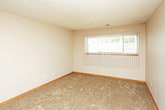 This single-floor unit presents a practical and well-organized layout across its living spaces. The main living area is finished with wall-to-wall carpet in a neutral beige tone, complemented by warm wood baseboards and a textured ceiling. A large window fitted with vertical blinds allows natural light to fill the room, creating a bright and open atmosphere. An entry door is accessible directly from this space, connecting to the adjacent areas.

The galley-style kitchen is efficiently designed with oak cabinetry lining both walls, providing generous storage above and below. Blue laminate countertops run the length of both sides, and the kitchen is equipped with a double stainless steel sink, a dishwasher, a full-size refrigerator, and an electric range. Light hardwood-style flooring runs throughout the kitchen, offering a clean contrast to the carpeted rooms.

Additional rooms in the unit continue the neutral color palette with beige walls, carpeted floors, and windows with blinds that invite natural light. The textured ceilings and consistent wood baseboards carry throughout, giving the unit a cohesive and uniform finish. The overall layout is straightforward, making efficient use of the available space.