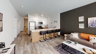 Located at 707 S Fourth St in Champaign, Illinois, this single-floor unit offers a thoughtfully designed layout with a clean, modern aesthetic throughout. The open-concept kitchen and living area serves as the heart of the space, featuring an extended island with bar seating, white upper cabinetry, warm wood lower cabinets, stainless steel appliances, and light wood-style flooring. A dedicated kitchen prep corridor provides additional counter space with matching cabinetry on both sides. The living area is bathed in natural light through large floor-to-ceiling windows with roller shades, creating a bright and welcoming atmosphere. Multiple bedrooms are well-proportioned, each offering large windows with views of the surrounding urban landscape, carpeted floors, and ample room for a bed and a dedicated study desk — ideal for student or professional living. The unit includes several bathrooms, each finished with warm wood vanities, white quartz countertops, framed mirrors, and glass-enclosed walk-in showers with white subway tile surrounds. Wood-look tile flooring carries through each bathroom, maintaining a consistent and polished design. The overall ambiance is contemporary, clean, and functional, blending modern finishes with practical living spaces.