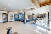 Located on District Lane in Chattanooga, Tennessee, this contemporary community clubhouse offers a thoughtfully designed, single-floor open-concept layout that blends comfort and entertainment seamlessly. The space is defined by light-toned luxury plank flooring, soaring ceilings with exposed spiral ductwork, and an abundance of natural light streaming through large windows and glass doors that open to an outdoor pool area.

The main gathering area serves as the heart of the space, anchored by a bold cobalt blue accent wall with a wall-mounted flat-screen TV and a stylish seating arrangement. A full-size billiards table positioned at the center makes for an ideal recreational hub. Structural columns clad in warm wood paneling add architectural character throughout.

A relaxed lounge nook near the entrance features hanging egg chairs and built-in bench seating set against natural wood-planked accent walls, creating an inviting retreat bathed in natural light. The kitchen area is sleekly appointed with white quartz countertops, gray cabinetry, a patterned tile backsplash, an under-counter beverage refrigerator, and pendant lighting, complemented by a communal bar-height dining table. Curated artwork and contemporary furnishings complete the modern aesthetic throughout this well-appointed community amenity space.