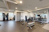 Located at 46180 Thomas Rd in Chilliwack, BC, this well-equipped fitness room offers a comprehensive workout facility within a compact, efficiently designed space. The room features durable rubber flooring throughout, recessed lighting, and a clean, modern aesthetic with white walls and a neutral color palette that creates a bright and motivating environment.

Large windows line one side of the room, allowing ample natural light to fill the space while offering views of the surrounding outdoor scenery. A wall-mounted flat-screen TV and a full-length mirror further enhance the workout experience.

The cardio zone is equipped with multiple Spirit treadmills and elliptical trainers, arranged to take advantage of the natural light from the windows. The strength training area features an Inspire multi-functional cable machine, a full dumbbell rack with a wide range of weights, a set of kettlebells, and adjustable workout benches. Wall-mounted resistance bands and boxing pads round out the available equipment, supporting a variety of training styles.

Exposed ventilation ducts on the ceiling add an industrial touch to the otherwise clean design. The space also includes a fire extinguisher, emergency signage, and key-card access, ensuring safety and security. Overall, this fitness room provides a well-rounded, functional environment suited for both cardio and strength training.