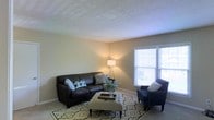 This single-floor space centers around a welcoming living room designed with a clean, neutral aesthetic. The room features warm beige walls and a textured ceiling that adds subtle visual interest overhead. Soft, carpeted flooring runs throughout, contributing to a comfortable and cozy atmosphere underfoot.

The living area is furnished with a dark leather sofa accented by decorative throw pillows, paired with a navy blue accent chair featuring nailhead trim detail. A tufted ottoman serves as a central coffee table, resting atop a patterned area rug with geometric motifs in muted tones of green and cream. A tripod floor lamp positioned in the corner provides warm ambient lighting that complements the natural light filtering in through the room's windows.

Two large windows with white horizontal blinds line one wall, allowing natural light to brighten the space and offer views to the exterior. The windows are framed in crisp white trim, providing a clean contrast against the soft wall color. A decorative centerpiece of greenery on the ottoman adds a touch of organic freshness to the room. The overall ambiance of the space is calm and inviting, with a balanced mix of functional furnishings and tasteful decor that creates a livable, well-composed interior.