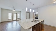This single-story residence is thoughtfully designed with an open-concept layout that connects the kitchen and living areas seamlessly. The kitchen serves as a central hub, anchored by a curved island with a light granite countertop and a double stainless steel sink. Three pendant lights hang above the island, providing focused illumination. Dark espresso cabinetry lines the kitchen walls, complemented by a mosaic tile backsplash, matching granite countertops, and black appliances including a range and built-in microwave. A stainless steel refrigerator adds contrast to the rich cabinetry. The adjacent living area features rich hardwood-style flooring, a ceiling fan, and a door providing direct outdoor access with natural light filtering through slatted blinds. Bedrooms are carpeted and well-lit by multiple windows with ceiling fans. The primary bathroom offers a double-sink vanity with dark cabinetry and granite countertops, dual framed mirrors, wood-style flooring, and access to a walk-in closet equipped with wire shelving and carpeted flooring. Throughout the space, neutral walls and consistent finishes create a cohesive and functional living environment.