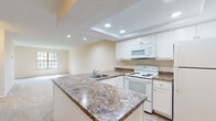 Located at 1363 Hideaway Woods Dr in Westerville, Ohio, this single-floor home offers a well-designed, open layout that balances functionality with comfort. The kitchen serves as a central focal point, featuring a large peninsula with striking granite countertops, a stainless steel sink, white cabinetry, and white appliances including a range, microwave, and refrigerator. Recessed lighting in a tray ceiling adds a refined touch to the kitchen area. Wood-look flooring runs through the kitchen and entry corridor, while plush carpet extends throughout the living areas and bedrooms, creating a warm, comfortable atmosphere. The expansive living room is bathed in natural light through large multi-pane windows and a glass-paneled door that opens to the outdoors, offering views of surrounding wooded scenery. Additional bedrooms are generously sized with carpeted floors, textured ceilings, and windows overlooking the natural landscape. One bedroom features a sliding glass door providing direct outdoor access. The bathroom is finished with a white vanity, wood-look flooring, and a full tub-shower combination. The overall color palette of soft cream and white tones throughout creates a bright, clean, and move-in-ready environment.