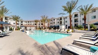 This expansive outdoor amenity area showcases a resort-inspired design that invites relaxation and social gatherings. At its heart is a generously sized swimming pool with crystal-clear turquoise water, surrounded by a broad concrete pool deck lined with lounge chairs ideal for sunbathing. Adjacent to the main pool, a bubbling spa sits beneath a shaded cabana area outfitted with striped blue curtains and comfortable seating, providing a private retreat from the sun. A striking outdoor fireplace serves as a focal architectural element near the spa, adding ambiance and a gathering point for cooler evenings. Tall, mature palm trees are planted throughout the space, lending a tropical, resort-like atmosphere and offering natural shade. The outdoor kitchen area is thoughtfully designed with a built-in stainless steel grill, prep sink, and storage, all set beneath a pergola with decorative tile accents — perfect for outdoor entertaining. Additional shade structures and umbrellas are distributed across the deck, complementing the sun-drenched environment. The multi-story residential buildings surrounding the courtyard feature a Mediterranean-inspired stucco exterior with red tile roofing and private balconies, creating an enclosed, community-focused setting that enhances the overall sense of privacy and luxury.