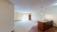 Located in Andover, Minnesota, this single-floor residence offers a thoughtfully designed layout that balances openness with functional living spaces. The main living area is generously proportioned, featuring plush carpeting and a sliding glass door that opens to a private balcony, allowing abundant natural light to pour in and creating a bright, airy atmosphere.

The kitchen is well-appointed with rich wood cabinetry, granite countertops, and black Whirlpool appliances including a range and over-the-range microwave. A raised breakfast bar with pendant lighting connects the kitchen to the living area, facilitating an open-concept flow. Hardwood flooring anchors the kitchen and entry zones, complementing the warm wood tones throughout.

Elegant French doors with frosted glass panes lead into a flexible secondary space, ideal as a bedroom, den, or office. The primary bedroom is carpeted, features a ceiling fan, and benefits from a window with outdoor views. Both bathrooms are equipped with accessibility features including grab bars, with one offering a pedestal sink and the other a granite-topped vanity with built-in cabinetry. In-unit laundry hookups are also present. Warm neutral tones and wood trim details lend a cohesive, inviting character throughout the entire space.