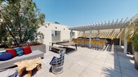 Located at 372 Fourth Ave in the heart of downtown San Diego, this rooftop terrace offers a well-appointed outdoor amenity space designed for both relaxation and recreation. The open-air deck is finished with large-format porcelain tile pavers that lend a clean, contemporary aesthetic throughout. A generous lounge area anchors one side of the terrace, furnished with a comfortable sofa accented with colorful throw pillows and complemented by a distinctive live-edge wood coffee table and modern wire accent chairs. A ping pong table provides a central recreational focal point. On the opposite end, a white pergola structure creates a shaded bar and dining zone, where bold yellow counter-height chairs line a sleek counter, and ornamental grasses in raised planters add a natural, textural element beneath the latticed canopy. Oversized modern planters throughout the deck feature mature olive trees, palms, and tropical shrubs, softening the urban setting. A perimeter railing opens to panoramic views of the San Diego skyline, allowing abundant natural light to fill the space. Elevator access ensures convenient entry to the terrace. The overall ambiance is modern and vibrant, reflecting the energy of its downtown San Diego location.