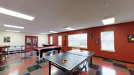 This single-floor recreational space is designed with leisure and social interaction in mind. The game room is highlighted by bold, deep-red accent walls that contrast with crisp white upper walls and ceiling, creating an energetic yet inviting atmosphere. The space is generously sized, comfortably accommodating multiple game stations without feeling crowded.

Two full-sized pool tables with red felt surfaces serve as focal points, positioned to allow ample movement around each. A ping-pong table is prominently featured in the foreground, complete with paddles and balls ready for play. A wall-mounted cue rack keeps billiard accessories organized and accessible.

Multiple windows along the walls allow natural light to fill the room, complementing the recessed panel lighting overhead that ensures consistent brightness throughout. The floor features a bold checkered tile pattern in red, black, and neutral tones, adding to the room's lively character.

Chess-themed wall art adds a thoughtful decorative touch, reinforcing the recreational theme. A glass-panel door at the far end provides access to an adjacent area, enhancing connectivity within the overall facility.