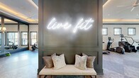 Located at 622 Brigham Rd in Greensboro, NC, this single-floor fitness center offers a generously sized, thoughtfully designed exercise space built to inspire and accommodate a wide range of workout routines. Upon entry, guests are greeted by a striking "live fit" neon sign mounted on a dark accent wall above a cushioned bench — setting a motivating tone from the moment you walk in.

The main exercise floor is expansive and open, featuring durable multi-tone flooring, tray ceilings with warm cove lighting, and ceiling fans throughout to maintain a comfortable environment. Large windows and floor-to-ceiling glass doors allow abundant natural light to flood the space, with views of the surrounding outdoor area including a pool courtyard.

The facility is equipped with a comprehensive lineup of strength and cardio equipment, including a multi-rack functional training system, free weights, adjustable benches, treadmills, stair climbers, ellipticals, rowing machines, and cable machines. Full-length mirrors line key sections of the walls, enhancing the sense of space and supporting form-focused training.

A dedicated turf sprint lane adds a performance training element, while elegant pendant lighting and sophisticated architectural details elevate the overall ambiance beyond a typical gym setting.