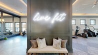 Located at 622 Brigham Rd in Greensboro, NC, this single-floor fitness center offers a generously sized, thoughtfully designed exercise space built to inspire and accommodate a wide range of workout routines. Upon entry, guests are greeted by a striking "live fit" neon sign mounted on a dark accent wall above a cushioned bench — setting a motivating tone from the moment you walk in.

The main exercise floor is expansive and open, featuring durable multi-tone flooring, tray ceilings with warm cove lighting, and ceiling fans throughout to maintain a comfortable environment. Large windows and floor-to-ceiling glass doors allow abundant natural light to flood the space, with views of the surrounding outdoor area including a pool courtyard.

The facility is equipped with a comprehensive lineup of strength and cardio equipment, including a multi-rack functional training system, free weights, adjustable benches, treadmills, stair climbers, ellipticals, rowing machines, and cable machines. Full-length mirrors line key sections of the walls, enhancing the sense of space and supporting form-focused training.

A dedicated turf sprint lane adds a performance training element, while elegant pendant lighting and sophisticated architectural details elevate the overall ambiance beyond a typical gym setting.