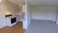 This single-floor residential unit offers a practical and functional layout centered around an open-concept design. The kitchen is equipped with white cabinetry, a built-in electric range, a dishwasher, and a sink, all set against light wood-style flooring that adds warmth to the space. A partial dividing wall with a built-in countertop bar conveniently separates the kitchen from the adjoining living area, creating a natural transition between the two spaces while maintaining an open feel. The living area features wall-to-wall carpet in a neutral tone, providing a comfortable and cohesive aesthetic. The textured ceiling runs throughout, and flush-mount lighting provides overhead illumination. The neutral color palette of white and gray tones throughout the unit creates a clean, understated ambiance that lends itself well to a variety of décor styles. The efficient use of space and the seamless flow between the kitchen and living area make this a well-organized and livable single-floor unit.