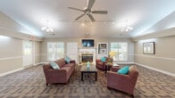 Located at 600 S Pine Hill Rd in Griffin, Georgia, this single-story community clubhouse offers a thoughtfully designed gathering space with a warm, inviting atmosphere. The centerpiece of the facility is a generously sized lounge area distinguished by soaring vaulted ceilings, a large ceiling fan, and dual chandelier fixtures that provide ample illumination. A classic white-framed fireplace serves as the focal point of the main seating area, accompanied by a wall-mounted flat-screen television and a built-in display shelf. Comfortable plush sofas and armchairs in rich mauve tones, accented with teal throw pillows, are arranged around dark wood coffee and side tables, creating an ideal setting for relaxed socializing. Multiple large windows fitted with white blinds allow abundant natural light to fill the room while offering views of the surrounding greenery. The patterned carpet throughout adds texture and visual interest to the space. Transitioning toward the rear of the clubhouse, a dining area features a dark wood table and chairs beneath a modern chandelier, positioned adjacent to a pass-through kitchen window. The kitchen, visible through the opening, includes cabinetry and countertop appliances. French doors at the back of the space provide additional access to the exterior, enhancing the open, connected feel of this well-appointed community facility.