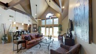 This single-story space is defined by its striking architectural design, most notably the dramatic vaulted ceilings that soar overhead, accented by exposed dark wood beams and ceiling fans that enhance the sense of grandeur. The open-concept layout connects the living area to an adjacent kitchen, creating a cohesive and functional flow throughout the space.

The living area serves as the focal point, anchored by a rich leather sofa with decorative pillows and complementary upholstered seating arranged around a glass-top coffee table. An ornate area rug grounds the seating arrangement, adding warmth and texture to the space. A large flat-screen television is mounted on the wall, flanked by decorative accents and lush potted plants that bring a natural element indoors.

A dramatic wall of windows, including a prominent arched transom window above a set of French doors, floods the space with natural light and provides views of the surrounding greenery. The warm, neutral wall tones complement the rich wood cabinetry visible in the kitchen, which features pendant lighting and stainless steel appliances. The overall ambiance is warm, inviting, and elegantly casual.