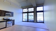 This contemporary urban space occupies a single floor and makes a strong architectural statement through its expansive floor-to-ceiling windows framed in bold black steel. These large-scale windows flood the interior with natural light while offering unobstructed views of the surrounding cityscape, including classic brick and stone urban buildings that speak to the historic character of the neighborhood.

The open main area features a combination of hardwood flooring and carpet, creating defined zones within the generous layout. Soaring ceilings add to the sense of spaciousness and volume, enhanced further by a suspended pendant light fixture that serves as a functional focal point. A track lighting system along the ceiling provides additional versatility for illuminating the space.

A dark-toned dresser or storage cabinet is positioned along one wall, complementing the neutral color palette of the walls and flooring. The overall design aesthetic blends modern industrial elements — evident in the steel window framing and clean architectural lines — with a light, airy ambiance created by the abundance of natural light.

The space presents a bright, open character well-suited for a variety of uses, with the dramatic city views serving as a defining and memorable feature throughout.