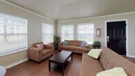 This single-floor office space serves as a functional and welcoming leasing or community hub, thoughtfully designed to accommodate both meetings and casual interactions. The layout is divided into two primary areas that flow seamlessly together, unified by warm hardwood-style flooring and crisp crown molding that runs throughout the space.

The meeting area is positioned near the entrance, featuring a bold black front door with a keypad entry system and a security camera overhead. A round dark-wood table with seating anchors this zone, while two large windows fitted with horizontal blinds allow abundant natural light to filter in, offering views of the surrounding greenery.

The adjacent lounge area is generously proportioned and equally well-lit, with multiple large windows that frame outdoor views and fill the room with daylight. Built-in window seat ledges add architectural character and provide additional surface space. Comfortable seating arrangements, including sofas and accent chairs, create a relaxed, inviting atmosphere well-suited for resident interaction. A prominent leasing banner adorns one wall, reinforcing the space's community-oriented purpose.

Throughout, the neutral warm-beige walls, white trim, and consistent flooring create a cohesive, professional aesthetic that feels both practical and approachable. The open, airy layout and abundance of natural light make this an especially functional and pleasant space.
