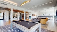 This thoughtfully designed single-floor amenity space in Tracy, California offers a dynamic blend of social and functional areas crafted with contemporary style. The open-plan layout is anchored by a spacious game room featuring a full-size pool table set atop a geometric patterned area rug, illuminated by a striking gold oval pendant light overhead. Light hardwood flooring flows throughout, unifying the various zones seamlessly.

Adjacent to the game room, a generously sized lounge area provides ample seating with a curved sectional sofa, accent chairs in bold yellow, and a sleek fireplace set within a floor-to-ceiling dark tile surround topped with a mounted television — creating an inviting gathering spot. Curated artwork adorns the white walls throughout, adding visual interest.

The kitchen area is a standout feature, showcasing marble-look countertops, a dramatic blue textured tile backsplash, white upper cabinetry with open shelving, and stainless steel appliances. Floor-to-ceiling windows flood the kitchen with natural light while offering views of an outdoor terrace equipped with professional-grade BBQ grills. Glass-partitioned private rooms are visible along one wall, adding flexible use space to the overall layout.