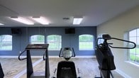 This single-floor fitness room is designed to provide a functional and comfortable workout environment. The space is defined by its deep navy-blue accent walls, which create a focused, energizing atmosphere complemented by light-toned wood-style flooring. Three arched windows line the primary wall, allowing generous natural light to fill the room while offering pleasant views of surrounding greenery, enhancing the overall sense of openness.

The room is equipped with a selection of cardio machines, including a treadmill, a stationary upright bike, and an elliptical or stair-climber, providing users with varied cardiovascular training options. A wall-mounted flat-screen television is positioned centrally above the windows, allowing exercisers to stay entertained during workouts. Flush panel ceiling lights ensure consistent, even illumination throughout the space.

A full-length mirror runs along one side wall, visually expanding the room and allowing users to monitor their form during exercise. The overall layout is clean and practical, with equipment arranged to allow comfortable movement between machines. The combination of natural light, mirrored surfaces, and modern finishes gives this fitness space a bright, welcoming character.