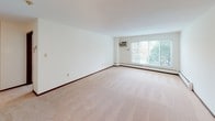 This single-floor residence offers a practical and comfortable layout across several well-defined living spaces. The open living area is generously proportioned, featuring plush wall-to-wall carpeting, crisp white walls with classic dark wood baseboards, and a large sliding glass window that invites abundant natural light and views of surrounding greenery. A wall-mounted air conditioning unit and baseboard heating provide year-round climate control. The adjacent dining and kitchen area continues the warm, functional aesthetic, with rich dark wood cabinetry offering ample storage, a built-in sink with countertop workspace, and a full-size refrigerator. A ceiling fan with integrated lighting keeps the space comfortable and well-lit. The bedrooms are carpeted and bright, each equipped with ceiling fans and windows overlooking outdoor foliage. One bedroom includes a spacious walk-in closet with built-in shelving, alongside an additional standard closet, providing excellent storage capacity. The full bathroom is cleanly finished with white tile surrounding a tub-shower combination, a mosaic tile floor, a toilet, and a dark wood vanity with a single sink and a mirrored medicine cabinet above. Throughout the unit, a consistent palette of neutral tones and natural wood accents creates a cohesive and inviting atmosphere.