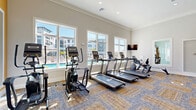 Located in Palmetto, FL, this well-appointed fitness center offers a comprehensive workout environment across two dedicated exercise rooms. The main exercise area is generously sized, featuring patterned carpet tiles in a blue and tan palette, crown molding, and recessed lighting complemented by ceiling fans. A bold golden-yellow accent wall energizes the space, while large mirrors visually expand the room. This area is equipped with a full suite of strength training machines, a rack with free weights and dumbbells, a Smith machine, and a cable system, catering to a wide range of resistance training needs. A wall-mounted television provides entertainment during workouts. The cardio zone showcases a row of treadmills, ellipticals, and a rowing machine positioned alongside large windows that frame views of the outdoor pool and surrounding community, flooding the room with abundant natural light. The second exercise room offers a more intimate setting with light wood-style flooring, soft blue-gray walls, and ample window coverage, making it ideal for cycling classes or stretching. Spin bikes and additional cardio equipment complete this bright, airy space. Throughout the facility, the design balances functionality with an inviting aesthetic, creating a motivating atmosphere for residents pursuing their fitness goals.