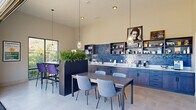 This contemporary two-floor space is thoughtfully designed for communal gathering and dining, offering a blend of modern aesthetics and functional layout across its two levels. The main floor serves as the heart of the space, anchored by a sleek kitchenette running along one wall, finished with striking blue square tile backsplash, dark wood lower cabinetry, white quartz countertops, and open upper shelving for display. Multiple dining configurations are arranged throughout, including a standard dining table with soft blue upholstered chairs and an elevated bar-height counter lined with bold purple and grey barstools — both illuminated by elegant geometric pendant lights. Tall ceilings amplify the sense of openness, while large windows and a full glass sliding door flood the interior with natural light and lush outdoor greenery views. Tall planter boxes with dramatic grasses serve as natural room dividers, adding life and texture to the interior. A glass door transitions seamlessly to the upper rooftop terrace — a generous open-air deck enclosed by sleek metal railings, furnished with woven lounge chairs, a dining table, and open views of the surrounding neighborhood beneath clear skies. Light-toned tile flooring runs consistently throughout both levels, tying the design together cohesively.