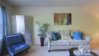 This two-story space offers a thoughtfully arranged living area characterized by a soft, neutral color palette that creates a calm and inviting atmosphere. The living room is anchored by a plush, light-colored sofa dressed with teal and blue accent pillows, complemented by a matching lounge chair that adds a pop of vibrant color to the room. A tall potted palm plant introduces a natural, organic element, enhancing the room's relaxed ambiance. A decorative folding screen panel adds a subtle textural contrast along one wall, contributing to a layered, curated aesthetic. Bold artwork depicting a peacock in full display serves as a striking focal point on the wall, injecting color and visual interest into the otherwise understated space. Soft carpeting underfoot adds warmth and comfort to the living area. The overall design balances neutral tones with strategic bursts of color, creating a space that feels both relaxed and visually engaging. Spanning two floors, the property offers versatile living arrangements suited to a variety of lifestyle needs.
