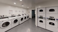 This shared laundry room is designed for efficiency and practicality, offering a clean, well-organized environment for residents. The space features multiple front-loading washing machines arranged in a row along one wall, each equipped with digital control panels for ease of use. Complementing the washers, stacked dryer units are positioned along the opposing wall, maximizing the available floor space while accommodating high-capacity laundry needs.

A built-in folding counter runs along one wall, providing a dedicated surface for sorting and folding laundry. The room is finished with durable speckled grey flooring that is both resilient and easy to maintain. Recessed lighting is installed throughout the ceiling, ensuring the space is consistently well-lit.

The walls are painted in crisp white and neutral grey tones, contributing to a clean and functional ambiance. Informational signage is posted on the walls, and a fire safety unit is visibly mounted near the entrance for compliance and safety. A louvered door provides access to an adjacent area. The overall design prioritizes utility, cleanliness, and ease of use, making it a functional shared amenity space.