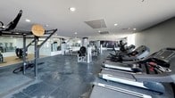 This fitness center is a thoughtfully designed, single-floor facility offering a comprehensive workout environment for users of all fitness levels. The space is characterized by its dark rubber tile flooring, which provides both durability and traction throughout the workout areas, and a clean white ceiling fitted with recessed lighting and integrated HVAC systems for consistent comfort.

The cardio zone features a row of treadmills and elliptical machines, providing ample options for aerobic training. Adjacent to this, a well-stocked strength training area houses multiple cable and resistance machines suited for a full-body workout. A dedicated free weights section includes a tiered dumbbell rack with a broad range of weights, while a power rack supports barbell and functional strength training.

Floor-to-ceiling mirrors line the walls throughout the space, visually expanding the interior and allowing users to monitor their form during exercise. A bold purple accent wall adds a dynamic visual contrast to the otherwise neutral palette, lending the facility a modern and energetic ambiance. Natural light filters in through windows, brightening the space and creating an open, welcoming atmosphere. The layout flows efficiently, with distinct zones for cardio, machine-based strength work, and free weight training, making this a versatile and fully equipped fitness destination.