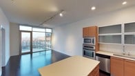 This single-floor space showcases a thoughtfully designed open-concept layout that seamlessly connects the kitchen and living areas. The kitchen is well-appointed with stainless steel appliances, including a built-in double wall oven and microwave, complemented by warm wood cabinetry and open shelving. A spacious island with a light-toned countertop anchors the kitchen, providing both prep space and a natural dividing point between the cooking and living zones. Track lighting runs along the ceiling, offering adjustable illumination throughout the space. Dark hardwood flooring adds a rich contrast to the bright white walls and ceiling, contributing to a clean, contemporary aesthetic. Floor-to-ceiling windows along one wall flood the interior with abundant natural light while framing expansive urban views, including a private balcony accessible through a glass sliding door. The open living area beyond the kitchen offers a versatile, airy atmosphere, making the most of the generous ceiling height. Overall, the space presents a modern, minimalist design with a strong emphasis on natural light and urban connectivity.
