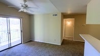 This single-floor space presents a straightforward and functional layout highlighted by neutral, beige-toned walls that create a calm and understated ambiance throughout. The main living area is finished with wall-to-wall carpeting in a muted tone, providing a comfortable underfoot feel and contributing to the room's understated character. A ceiling fan is mounted overhead, offering both air circulation and modest overhead lighting to the space.

A centrally positioned HVAC vent on the wall indicates a central air system, ensuring climate control throughout. The room features standard white baseboards that cleanly frame the carpeted floor, adding a subtle finishing detail to the walls.

A doorway visible from the main living area leads into a hallway, illuminated by a wall-mounted light fixture, suggesting access to additional rooms beyond. The hallway continues the same neutral color palette and carpeted flooring, maintaining visual consistency across the space.

Overall, the space presents a clean, no-frills interior that is move-in ready and well-suited for personalization. Its simple design and neutral finishes provide a versatile backdrop adaptable to a variety of furnishing styles and décor preferences.