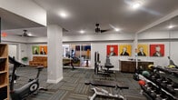 Located at 1525-1541 N High St in Columbus, Ohio, this fitness center offers a thoughtfully designed, single-floor space dedicated to health and wellness. The layout is open and functional, providing ample room for a diverse range of exercise equipment without feeling cramped.

The cardio zone features multiple treadmills and elliptical machines from True Fitness, each positioned in front of wall-mounted flat-screen televisions for an engaging workout experience. A large floor-to-ceiling mirror spans one wall, enhancing the sense of space and allowing users to monitor their form. Nearby, a well-stocked free weight area includes a full rack of dumbbells and medicine balls for versatile strength training.

The strength and functional training section houses a multi-station cable machine, a rowing machine, and a weight bench, catering to a wide variety of workout routines. A wooden cubby storage unit provides convenient organization for personal items or accessories. An accent wall finished in a deep teal textured tile adds a striking visual element, complementing the crisp white walls throughout.

The flooring consists of multi-toned striped carpet tiles in shades of blue, gray, and green, adding color and texture underfoot. Recessed lighting and a ceiling fan ensure the space remains well-lit and comfortable. Glass doors and interior windows connect the fitness center to adjacent common areas, contributing to an open, community-oriented atmosphere.