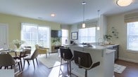This single-floor space showcases an open-concept layout that integrates the kitchen and living area into one cohesive, light-filled environment. The kitchen features a sleek white peninsula with seating for multiple bar-height stools, making it an ideal spot for casual dining or entertaining. Pendant lighting hangs above the peninsula, providing focused illumination that complements the natural light streaming through multiple windows fitted with Roman shades. The kitchen is equipped with a modern sink and ample counter space, maintaining a clean, minimalist aesthetic.

The adjoining living area is warm and inviting, anchored by rich hardwood-style flooring that flows throughout the space. Large sliding glass doors open to an outdoor area, flooding the interior with daylight and offering views of the surrounding greenery. The living space accommodates comfortable seating and is finished with soft, neutral-toned walls that enhance the airy atmosphere. A wall-mounted television serves as a focal point. Together, the rooms create a functional and welcoming environment well-suited for both everyday living and entertaining.