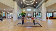 This single-story clubhouse is designed to impress, anchored by a sweeping grand lobby with soaring vaulted ceilings clad in dark wood planking. Prominent limestone columns frame the open space, while a dramatic circular chandelier with layered string lighting serves as a striking focal point overhead. Wide-plank hardwood flooring flows throughout, and clerestory windows allow natural light to fill the voluminous interior. A live-edge wood console table centered on a bordered area rug sets an elegant yet approachable tone, flanked by curated seating arrangements in rich jewel tones.

The space extends into multiple distinct zones. A lounge area with large windows offers views of an outdoor pool and courtyard, featuring a shuffleboard table and access through glass-paneled barn-style doors. A dedicated coffee bar area showcases gray shaker cabinetry, subway tile backsplash, a stainless steel refrigerator, quartz countertops, and a seated peninsula illuminated by brass pendant lights—ideal for casual gathering.

The fitness center is well-appointed with cardio equipment, a cable machine, free weights, and plyometric boxes, all set beneath a tray ceiling with accent lighting and a ceiling fan. Natural light enters through exterior-facing windows, and glass-paneled doors connect the gym directly to the outdoor amenity area.
