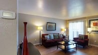 This single-floor space offers a welcoming and functional layout designed for comfortable everyday living. The living area serves as the heart of the space, featuring warm hardwood-style flooring that adds a touch of natural elegance throughout. A textured ceiling lends subtle depth to the room, while neutral-toned walls provide a versatile backdrop that complements a variety of décor styles. The living room is well-proportioned, offering ample space for seating and relaxation. A plush, deep-red sofa anchors the room and is accented with decorative teal and patterned throw pillows, adding a pop of color and character. A sleek wooden coffee table sits at the center, providing both style and functionality. Ambient lighting is thoughtfully provided through floor lamps and table lamps, creating a warm, inviting atmosphere throughout the day and evening. A large window fitted with vertical blinds allows natural light to filter into the space, brightening the interior and offering a connection to the outdoors. Framed wall art adds a personal, curated touch to the living area. Overall, the space balances comfort and practicality, making it an ideal setting for both relaxing and entertaining.