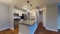 Located in Auburn Hills, Michigan, this single-floor residence offers a thoughtfully designed layout with modern finishes and a cohesive aesthetic throughout. The open-concept kitchen is a standout feature, boasting crisp white shaker cabinets, granite countertops, and a central island with a built-in sink and pendant lighting overhead. Stainless steel appliances, including a range, microwave, refrigerator, and dishwasher, complete the fully equipped kitchen, which flows seamlessly into the adjacent dining and living areas. Rich, dark hardwood-style flooring runs throughout the main living spaces, adding warmth and continuity to the interior. The living room is generously proportioned, with large windows that invite natural light and views of the surrounding exterior. Bedrooms are well-sized, featuring the same hardwood-style floors, neutral gray walls, and ample natural light from window treatments. The bathroom is finished with granite countertops on a white vanity, large-format stone-look tile flooring, and a tub-shower combination with marble-style surround. A dedicated laundry room accommodates a full-size washer and dryer with overhead shelving for additional storage. The overall ambiance is clean, modern, and move-in ready.