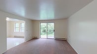 This single-floor unit offers a practical and well-organized layout designed for comfortable everyday living. The generously sized living room is carpeted in a neutral tone and filled with natural light, featuring a sliding glass door that opens to lush, tree-lined outdoor views and is equipped with baseboard heating. An adjoining room, visible through an open passageway, provides additional flexible living or dining space with its own window and baseboard heater.

The kitchen is efficiently designed with warm wooden cabinetry, white countertops, and a peninsula with a breakfast bar overhang. A white refrigerator, built-in sink, and louvered pantry doors add to its functionality, while light-toned flooring ties the space together.

Two carpeted bedrooms offer quiet retreats, each featuring windows that bring in natural light and baseboard heating for year-round comfort. The rooms are finished with neutral walls, providing a clean, versatile canvas.

The bathroom includes a wide vanity with wooden cabinetry, a light-toned countertop, a large mirror, and tile-style flooring. The space is straightforward and functional, with ample storage beneath the sink. Overall, the unit presents a clean, move-in ready layout with consistent finishes throughout.