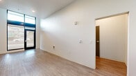 Located on North Vancouver Avenue in Portland, Oregon, this thoughtfully designed single-floor unit offers a clean, modern aesthetic throughout its compact footprint. The space is organized across three distinct areas, each finished with warm light wood-look flooring and crisp white walls that create a bright, cohesive atmosphere.

The main living area benefits from dramatic floor-to-ceiling windows and a glass entry door framed in sleek black, allowing abundant natural light to pour in and providing views of the surrounding streetscape. Soaring ceiling height in this zone further enhances the sense of openness. Recessed lighting throughout supplements natural light, maintaining a well-lit environment at all times.

The kitchen is efficiently designed with a continuous run of two-tone cabinetry — grey lower cabinets and white upper cabinets — paired with a white countertop. It includes an integrated cooktop, a single-basin sink with a modern gooseneck faucet, a built-in microwave, and a full-size refrigerator. Open upper shelving adds functional display space, while a window above the counter brings in additional daylight.

A separate room with recessed lighting and clean white walls provides a versatile private space, suitable for use as a bedroom or home office. The overall design is minimal and contemporary, making efficient use of every square foot while maintaining a fresh, livable ambiance.