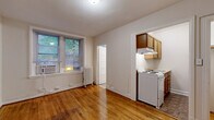 This single-floor unit offers a functional and straightforward layout across several distinct areas. The main living space is characterized by warm, honey-toned hardwood floors that extend throughout, neutral white walls, and natural light streaming in through windows fitted with blinds and a window air conditioning unit. A classic cast-iron radiator provides traditional heating, adding a touch of timeless character to the room.

The kitchen is a separate, defined space accessible directly from the main living area. It features warm wood cabinetry, a white electric range with an overhead ventilation hood, and a sink, offering all essential cooking amenities in a compact footprint. The flooring transitions to a durable tile surface within the kitchen.

A dedicated closet area with a hanging rod and coat hooks is conveniently situated near the entryway, providing practical storage. The entry itself includes a solid wood door and an additional white panel door leading to the closet.

The bathroom is a full, self-contained space accessed via a paneled door. It includes a toilet, a white vanity with sink and medicine cabinet above, a bathtub, and a vanity light fixture. The floor is covered in earth-toned tile, giving the room a clean, neutral appearance.

Overall, the unit maintains a clean, minimalist aesthetic with classic architectural details, making it a practical and well-organized living space.
