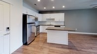 This single-floor space offers a thoughtfully designed layout with modern finishes throughout. The kitchen serves as a central highlight, featuring crisp white cabinetry, a classic subway tile backsplash, dark countertops, and a full suite of stainless steel appliances including a refrigerator, range, microwave, and dishwasher. A central island provides additional prep and dining space, while warm wood-tone flooring ties the area together and extends toward the entryway. Recessed lighting illuminates the kitchen and living areas, creating a bright and functional environment. The bedrooms are finished with plush wall-to-wall carpeting and neutral-toned walls, offering a comfortable and quiet retreat. One bedroom includes a ceiling fan and a generously sized window that invites natural light and offers open views of the surrounding cityscape. The bathroom is clean and functional, with wood-look tile flooring, a white vanity with dark countertops, a large mirror, and a combined shower and tub. The overall design aesthetic is contemporary and cohesive, with a neutral color palette of grays, whites, and warm wood tones that lend the space a fresh, move-in-ready appeal.