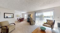 This single-floor residence offers a comfortable and functional layout designed for everyday living. The open-plan living and dining area is bright and welcoming, featuring plush wall-to-wall carpeting, a textured ceiling, and large sliding glass doors fitted with vertical blinds that lead to a private balcony overlooking lush outdoor scenery. The living room provides generous space for seating and entertainment, complete with open shelving for media and décor. Adjacent to the living area, the dedicated dining space accommodates a full dining set and receives ample natural light through a window with blinds.

The galley-style kitchen is efficiently arranged with warm wood cabinetry, laminate countertops, a double sink, and durable wood-look flooring. It includes essential appliances and offers practical storage throughout.

The primary bedroom is a restful retreat with soft carpeting, good natural light, and direct access to an en-suite bathroom. The second bedroom is equally comfortable, featuring a bright window dressed with curtains and blinds. Both bathrooms are clean and functional — one featuring a tub-shower combination and wood-look flooring, and the other a compact vanity setup with white fixtures. Together, the rooms create a cohesive, livable space with a casual, coastal-inspired aesthetic.