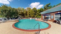 Located at 221 Fairforest Way in Greenville, South Carolina, this outdoor amenity space centers around a generously sized freeform swimming pool with classic brick coping and a stamped concrete surround. The pool features built-in water jets and fountains that add a resort-like ambiance, along with multiple entry points via stainless steel handrails and a shallow wading area. The expansive pool deck is outfitted with rows of lounge chairs ideal for sunbathing, along with multiple shaded seating areas furnished with tables and chairs beneath blue market umbrellas. A brick-accented clubhouse structure flanks one side of the pool area, offering additional covered patio space for relaxing or socializing. The surrounding landscape is lush with mature trees that provide natural shade and a sense of privacy. A wrought-iron perimeter fence encloses the space, ensuring security while maintaining open sightlines to the greenery beyond. Multi-story residential buildings with traditional architectural detailing, including pitched rooflines and chimneys, frame the backdrop of the pool area, creating a cohesive and inviting community atmosphere. The space is well-suited for warm-weather recreation and outdoor gatherings.