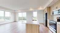 This thoughtfully designed 3-bedroom, 1-bathroom unit offers a clean, contemporary aesthetic with light wood tones, neutral walls, and an abundance of natural light throughout. The open-concept kitchen and living area serves as the heart of the home, anchored by a large center island with a white quartz countertop and an undermount sink. Light wood cabinetry pairs seamlessly with stainless steel appliances, including a refrigerator, electric range, and built-in microwave. Recessed lighting and sliding glass doors leading to a private balcony with green treetop views complete the bright, airy living space. The three bedrooms are finished with plush carpet flooring, and large windows offer elevated outdoor views of the surrounding area. The spacious primary bedroom includes a generous walk-in closet fitted with wire shelving. A well-appointed full bathroom features a dual-sink vanity with a quartz countertop, light wood cabinetry with matte black hardware, a large framed mirror, and a glass-enclosed walk-in shower with white subway tile. A clean hallway with modern doors and light wood flooring connects all areas of the unit, creating a cohesive and functional layout throughout.