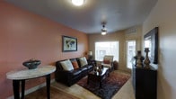 This single-floor residence offers a comfortable and cohesive living environment designed with both function and style in mind. The living room serves as the centerpiece of the space, immediately drawing attention with its rich terracotta-toned accent wall that adds warmth and character to the interior. Light-colored tile flooring runs throughout, providing a clean and durable foundation that complements the overall color palette. A ceiling fan with an integrated light fixture occupies the center of the room, ensuring both air circulation and ambient illumination. Natural light flows generously into the living area through a large window fitted with white blinds, alongside a glass-paneled exterior door that further brightens the space and offers outdoor access. The room comfortably accommodates a dark leather sofa adorned with decorative pillows, a matching armchair, and a dark wood coffee table set atop a layered area rug, creating a defined and inviting seating arrangement. A framed world map print adds a global, worldly aesthetic to the accent wall. The layout is efficient and well-proportioned, with each element contributing to a cohesive, lived-in atmosphere that is both practical and visually appealing.