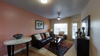 This single-floor residence offers a comfortable and cohesive living environment designed with both function and style in mind. The living room serves as the centerpiece of the space, immediately drawing attention with its rich terracotta-toned accent wall that adds warmth and character to the interior. Light-colored tile flooring runs throughout, providing a clean and durable foundation that complements the overall color palette. A ceiling fan with an integrated light fixture occupies the center of the room, ensuring both air circulation and ambient illumination. Natural light flows generously into the living area through a large window fitted with white blinds, alongside a glass-paneled exterior door that further brightens the space and offers outdoor access. The room comfortably accommodates a dark leather sofa adorned with decorative pillows, a matching armchair, and a dark wood coffee table set atop a layered area rug, creating a defined and inviting seating arrangement. A framed world map print adds a global, worldly aesthetic to the accent wall. The layout is efficient and well-proportioned, with each element contributing to a cohesive, lived-in atmosphere that is both practical and visually appealing.