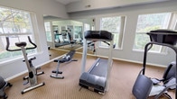 This fitness center offers a bright and functional workout environment designed to accommodate a variety of exercise needs. The space features vaulted ceilings that contribute to an open, airy feel, while ceiling fans provide comfortable air circulation throughout. Neutral gray walls and carpeted flooring create a clean, understated aesthetic throughout.

The workout area is equipped with several cardio machines, including treadmills, an elliptical trainer, and a stationary bike, alongside free weights, a weight bench, and a stability ball. A large wall-mounted mirror spans one side of the room, effectively amplifying the sense of space and allowing users to monitor their form during exercise.

Natural light filters in through multiple windows positioned along the walls, offering views of surrounding greenery and keeping the environment feeling fresh and inviting. A dedicated storage credenza in warm wood tones provides convenient space for accessories or personal items.

A pair of elegant French doors with gold hardware connects the fitness center to an adjacent lounge or common area, where hardwood flooring, colorful seating, and additional windows are visible, creating a seamless transition between spaces. A direct exterior door also provides convenient outdoor access. Together, these elements make this fitness center a practical and pleasant space for daily exercise.