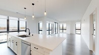 This well-appointed 3-bedroom, 2-bathroom residence offers a thoughtfully designed single-floor layout with a clean, contemporary aesthetic. The open-concept living and kitchen area serves as the heart of the home, bathed in natural light from expansive floor-to-ceiling windows with sleek black frames that frame outdoor views. The kitchen is a standout feature, boasting white shaker cabinetry with matte black hardware, a large quartz waterfall island with an undermount sink, pendant lighting, and stainless steel appliances. It flows seamlessly into a spacious living area finished with wood-look plank flooring and access to an outdoor balcony. The three bedrooms are carpeted for comfort, each featuring large windows and generous closet space, including walk-in closets with built-in hanging rods. The primary bedroom offers a notably spacious footprint. Both bathrooms are finished in a bright, neutral palette; one features a glass-enclosed walk-in shower with white tile surround, light tile flooring, and a white vanity with quartz countertop. Throughout the residence, crisp white walls, consistent modern finishes, and abundant natural light create a bright, airy ambiance that is both functional and inviting.