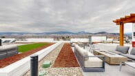 This impressive rooftop terrace, located at 500 Asher Rd in Kelowna, British Columbia, offers a multifaceted outdoor experience with breathtaking panoramic views of the surrounding mountains. The space is thoughtfully divided into distinct areas, each serving a unique purpose. A large artificial grass area provides a lush, green space ideal for various activities. Adjacent to this is a contemporary lounge area furnished with sleek, modern outdoor seating, perfect for relaxation and socializing. The terrace also incorporates eco-friendly elements, including areas of drought-resistant plantings and decorative stone sections, demonstrating a commitment to sustainable design. A covered pergola area offers shade and additional seating options. The terrace is bordered by glass railings, ensuring unobstructed views while maintaining safety. Ample lighting fixtures are strategically placed throughout, allowing for enjoyment of the space well into the evening. The clean lines of the white exterior walls and grey tiled flooring contribute to the overall modern aesthetic. This well-designed rooftop oasis provides an ideal setting for both private relaxation and entertaining, all while showcasing the natural beauty of Kelowna's landscape.