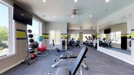 This fitness center offers a comprehensive workout environment designed to accommodate a wide range of exercise needs. The space is finished with a patterned carpet in blue and yellow tones, complemented by bold horizontal stripes of green, yellow, and blue that run along the neutral gray walls, creating an energetic and motivating atmosphere. Recessed lighting and ceiling-mounted fans ensure the space remains well-lit and comfortable during workouts. Large windows along multiple walls flood the interior with natural light and offer views of the surrounding greenery, enhancing the open and airy feel of the room. The cardio section is well-stocked with treadmills, stationary bikes, and a rowing machine, while a dedicated free weights area features a medicine ball rack with a variety of weighted balls and exercise balls. An adjustable weight bench provides space for strength training exercises. A wall-mounted flat-screen television is positioned for easy viewing during workouts. A full-length mirror along one wall helps users monitor their form, and a water station is conveniently accessible. The layout is thoughtfully organized to allow ample circulation space between equipment, making the fitness center both functional and inviting.
